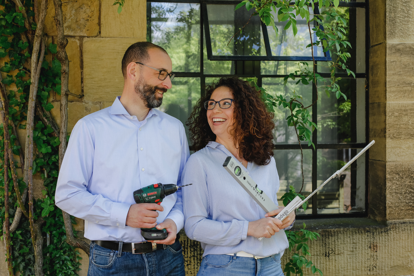 Zwei Personen stehen vor einem Fenster, der Mann hält eine Bohrmaschine und die Frau ein Lineal.